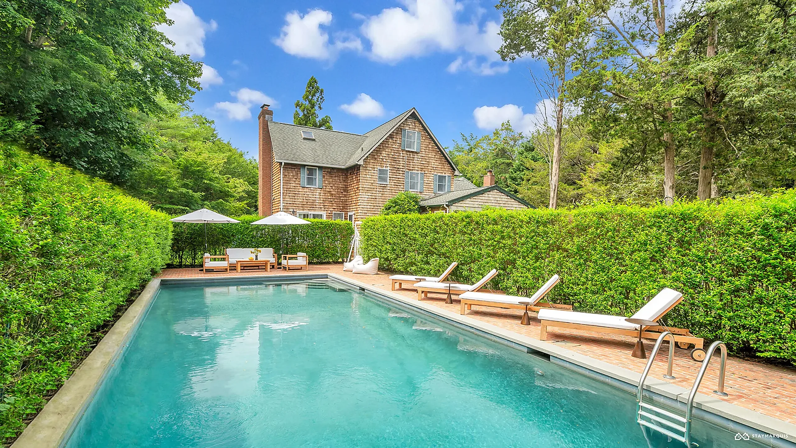 Cedar-shingle vacation home behind tall clipped hedges, with a long turquoise pool and teak sun loungers